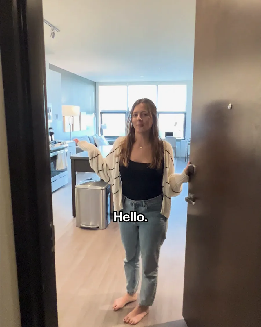 A video still: a woman in a striped cardigan holds open an apartment door and welcomes the viewer in, the caption "Hello." on screen.