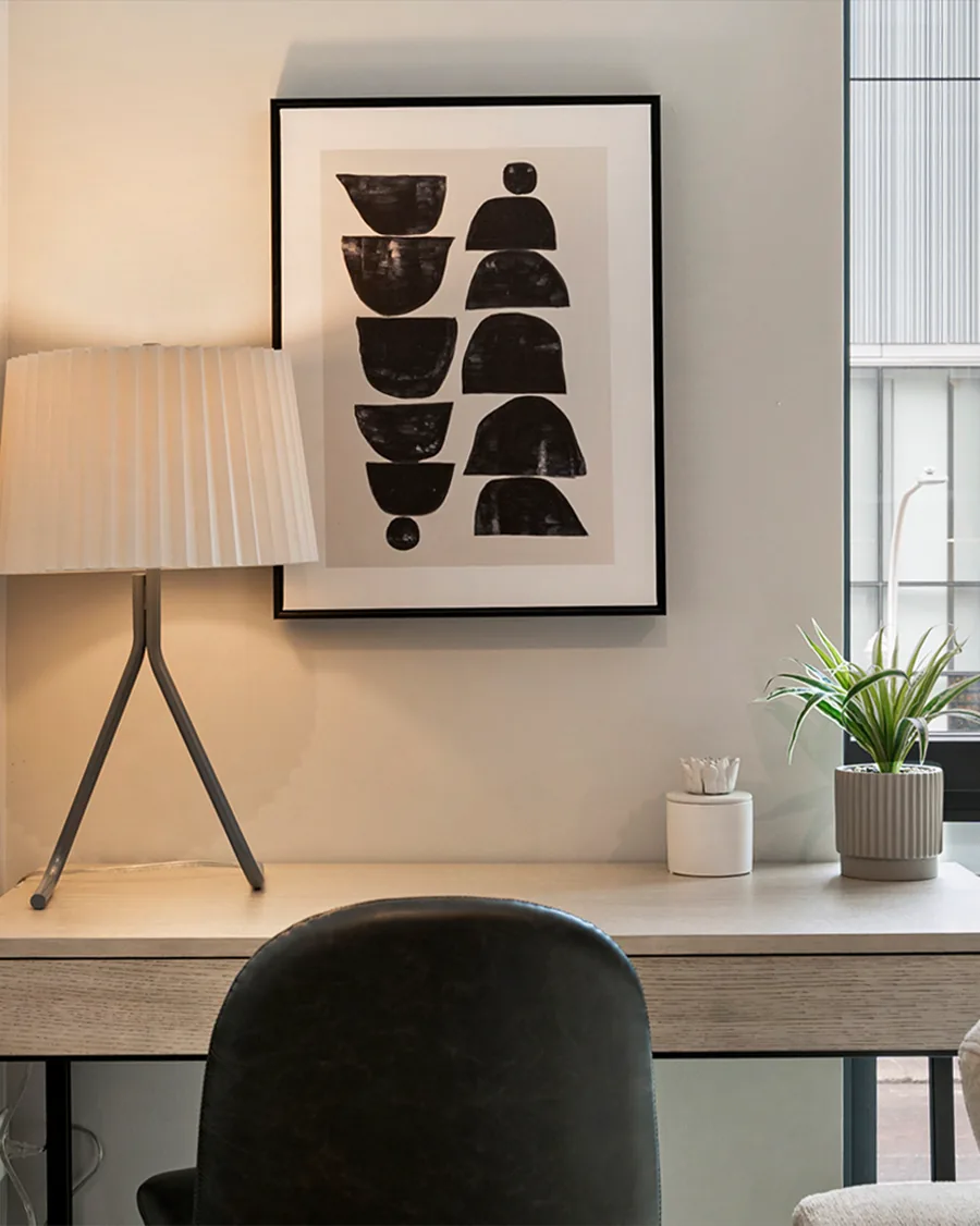 A tall photo of a work nook: a pleated lamp and a framed black and white print over a pale desk with a dark chair.