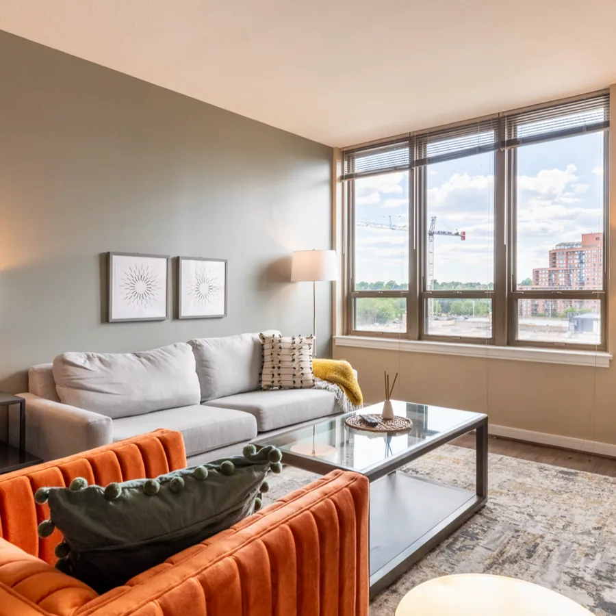 A square photo of a living room: a grey sofa, a glass coffee table and two orange velvet chairs below wide windows over a city.