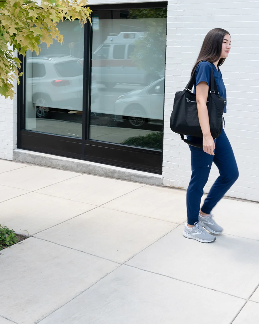 A tall photo: a nurse in navy scrubs with a black tote strides along a white brick wall, an ambulance reflected in the window.