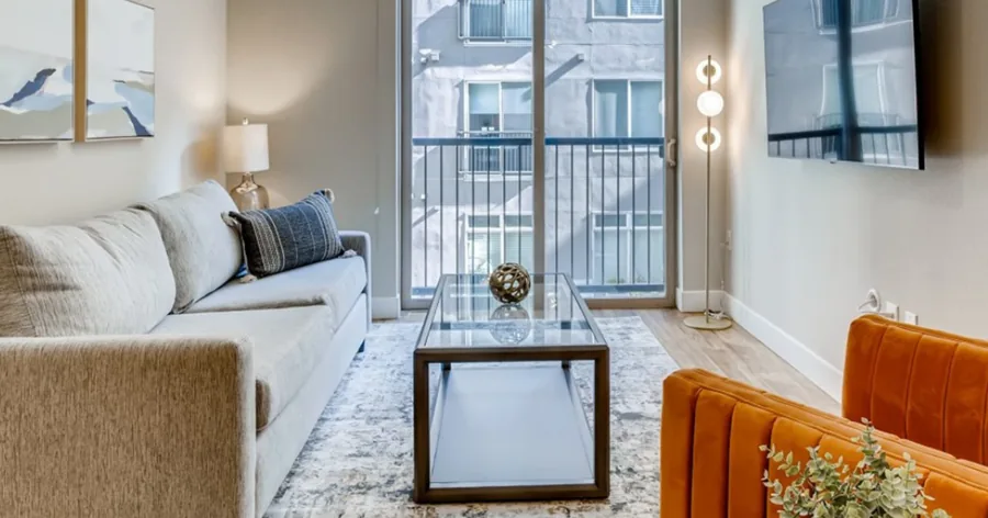 A wide photo of a furnished living room: a grey sofa, a glass coffee table, an orange chair and a balcony door with city light behind.