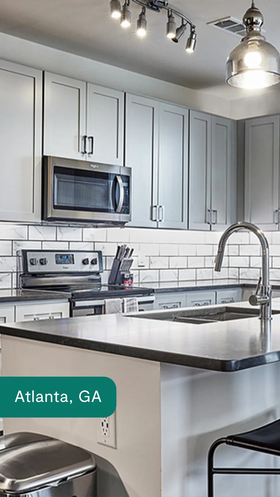 A tall ad: a grey kitchen with pendant and track lights, a dark stone island and a steel sink, the city's name on a green tab at the bottom.