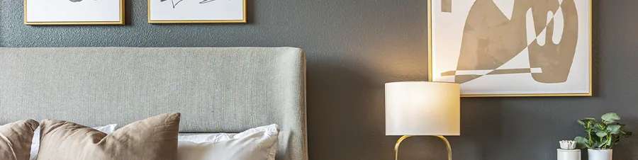 A wide strip of a bedroom: a padded headboard, a warm lamp and framed abstract prints on a dark wall.