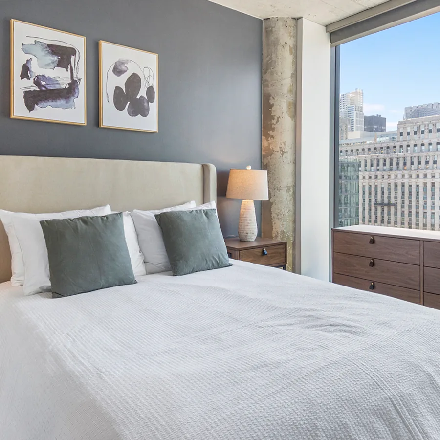 A square photo of a bedroom: a white bed with sage pillows, two abstract prints on a slate wall, and a window over tall city buildings.