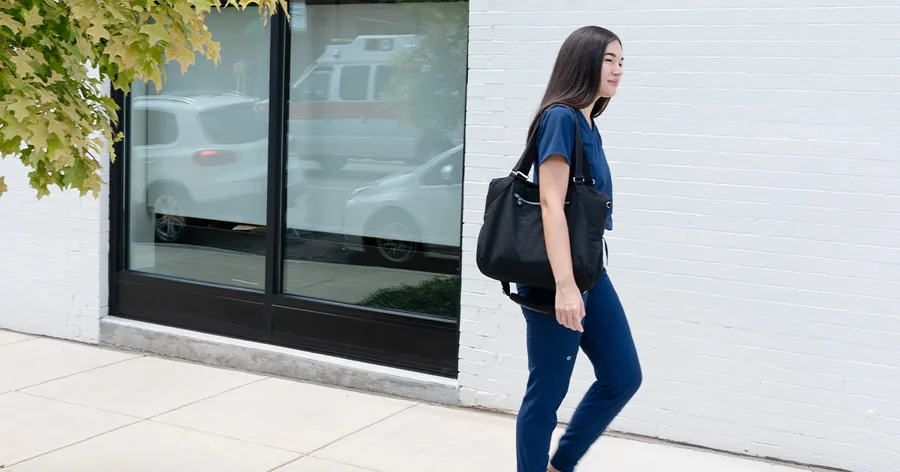 A wide photo: a nurse in navy scrubs with a black tote walks past a white brick wall and a window reflecting an ambulance.