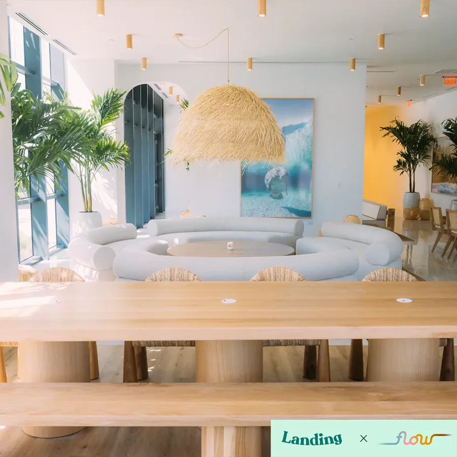 A square photo of a bright shared lounge: a long wood table, a round white sofa, palms and a woven pendant lamp, two logos in the corner.