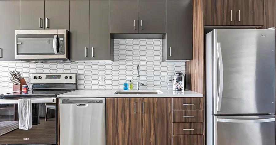 A wide photo of an apartment kitchen: dark wood cabinets, a white tile backsplash and steel appliances.