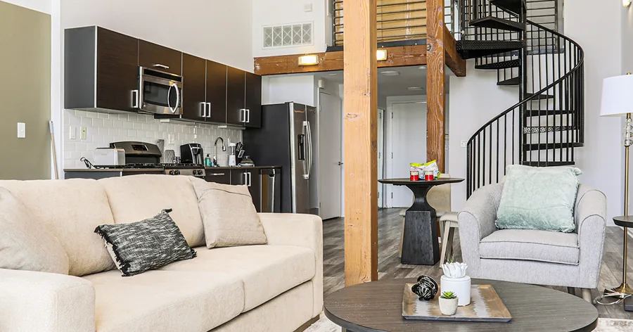 A wide photo of a loft apartment: a cream sofa, a round coffee table, a dark kitchen and a black spiral staircase behind a timber post.