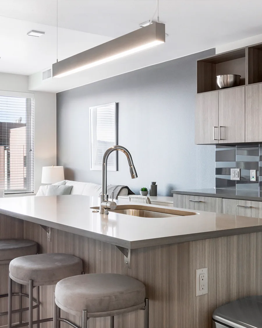 A tall photo of an apartment kitchen: a pale counter with a steel sink and round stools under a long hanging light.