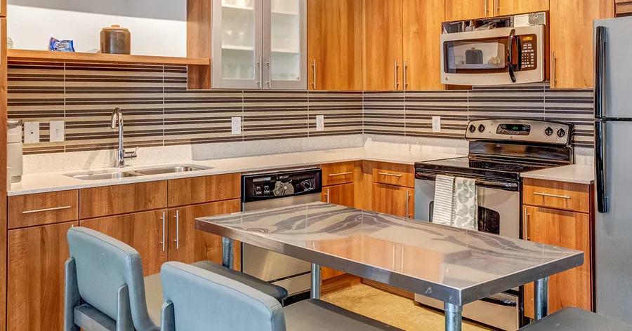 A wide photo of an apartment kitchen: wood cabinets, a striped tile backsplash, steel appliances and a steel island with blue stools.