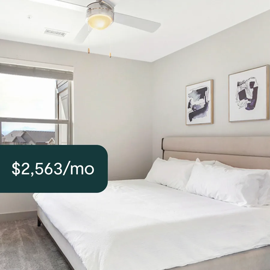 A square ad: a bright bedroom with a padded headboard, white bedding and two framed prints, a dark tab with the monthly rent.