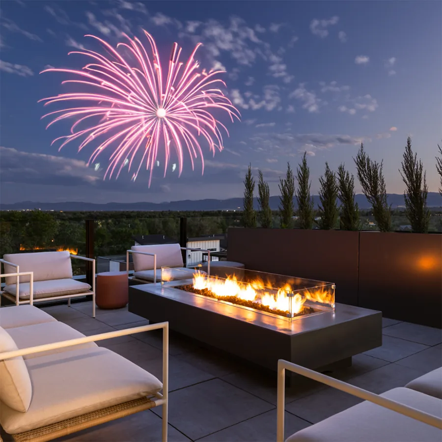 A square photo of a rooftop lounge at dusk: white sofas around a long fire table, pink fireworks bursting in the sky above.