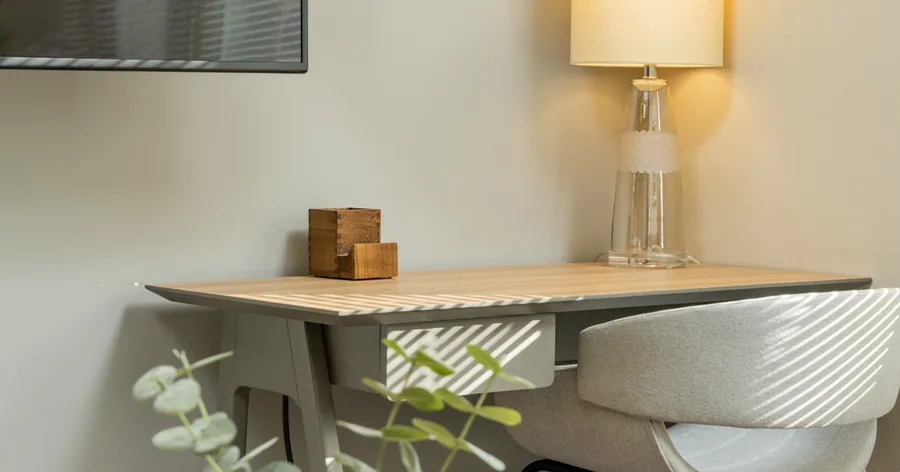 A wide photo: a small wood desk and a white chair under a glass table lamp, a sprig of eucalyptus in the foreground.