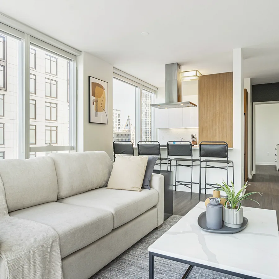 A square photo of a bright corner apartment: a cream sofa, a marble coffee table and a kitchen counter with black stools against tall windows.