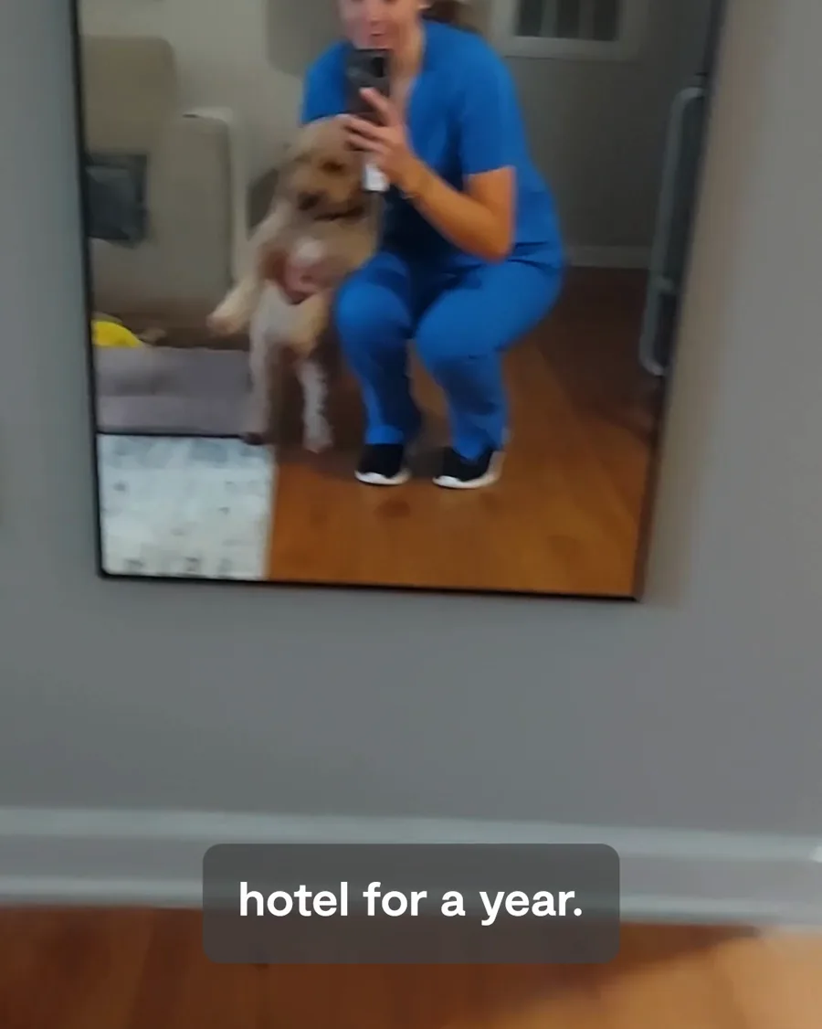A video still: a nurse in blue scrubs holds a fluffy dog in a mirror selfie, the caption "hotel for a year." along the bottom.