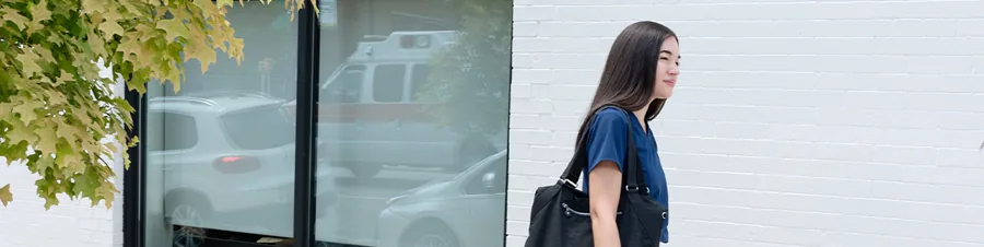 A thin banner crop: a nurse in navy scrubs walks past a white brick wall, leaves in the corner.