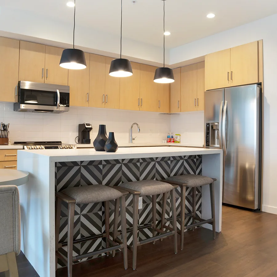 A square photo of an apartment kitchen: three black pendant lamps over a white island with a chevron tile front and three stools.