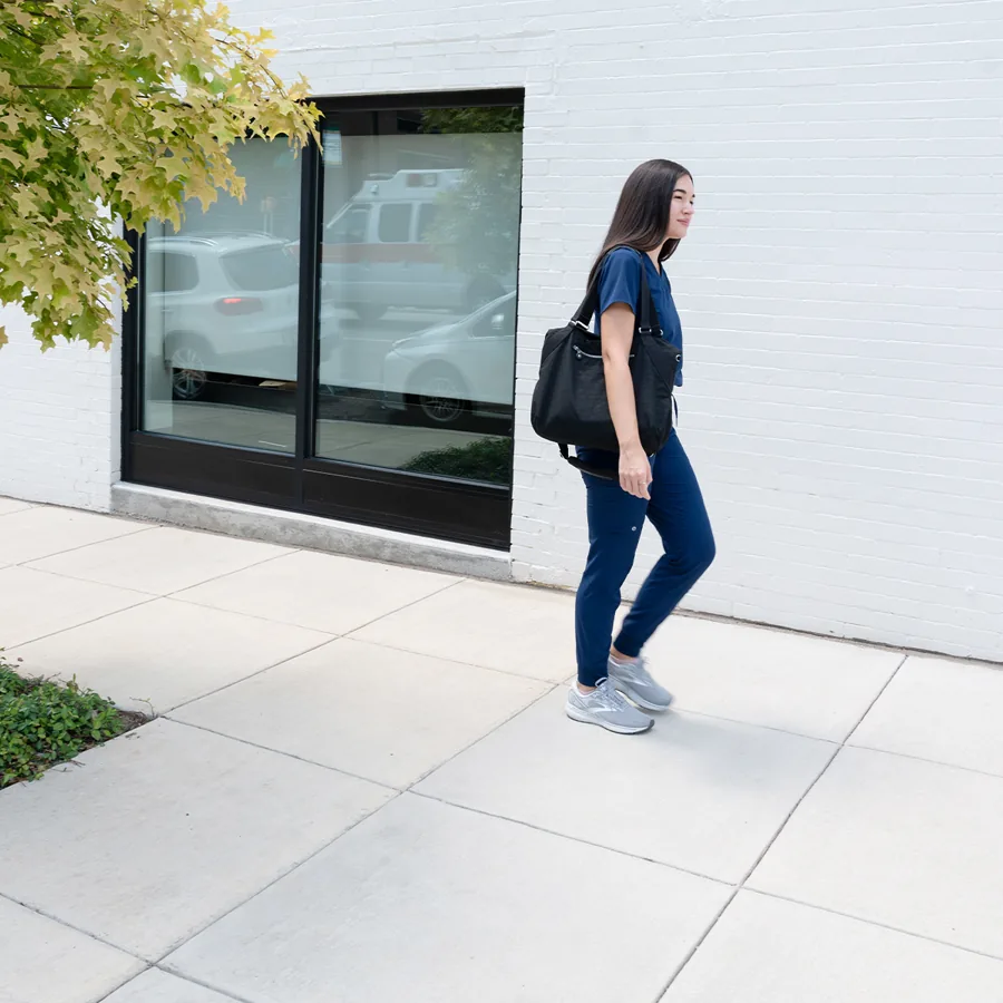 A square photo: a nurse in navy scrubs and grey sneakers walks a pale sidewalk past a white brick wall and dark window.